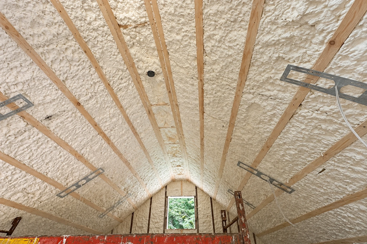 White spray foam covers the sloped rafters of a tall attic, with exposed wooden beams and a small window.