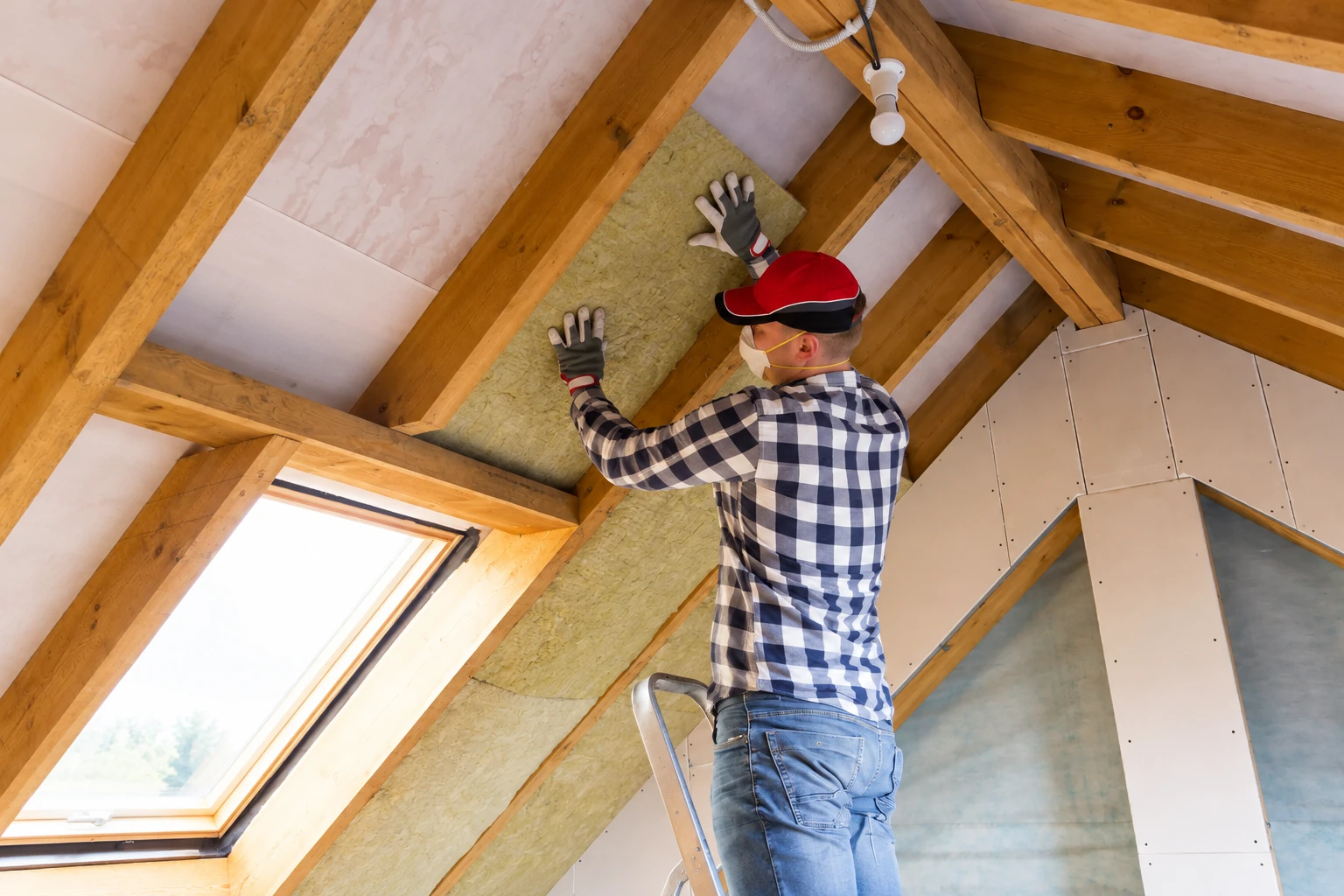 A worker in gloves and a mask fits yellow batt insulation between exposed attic rafters.