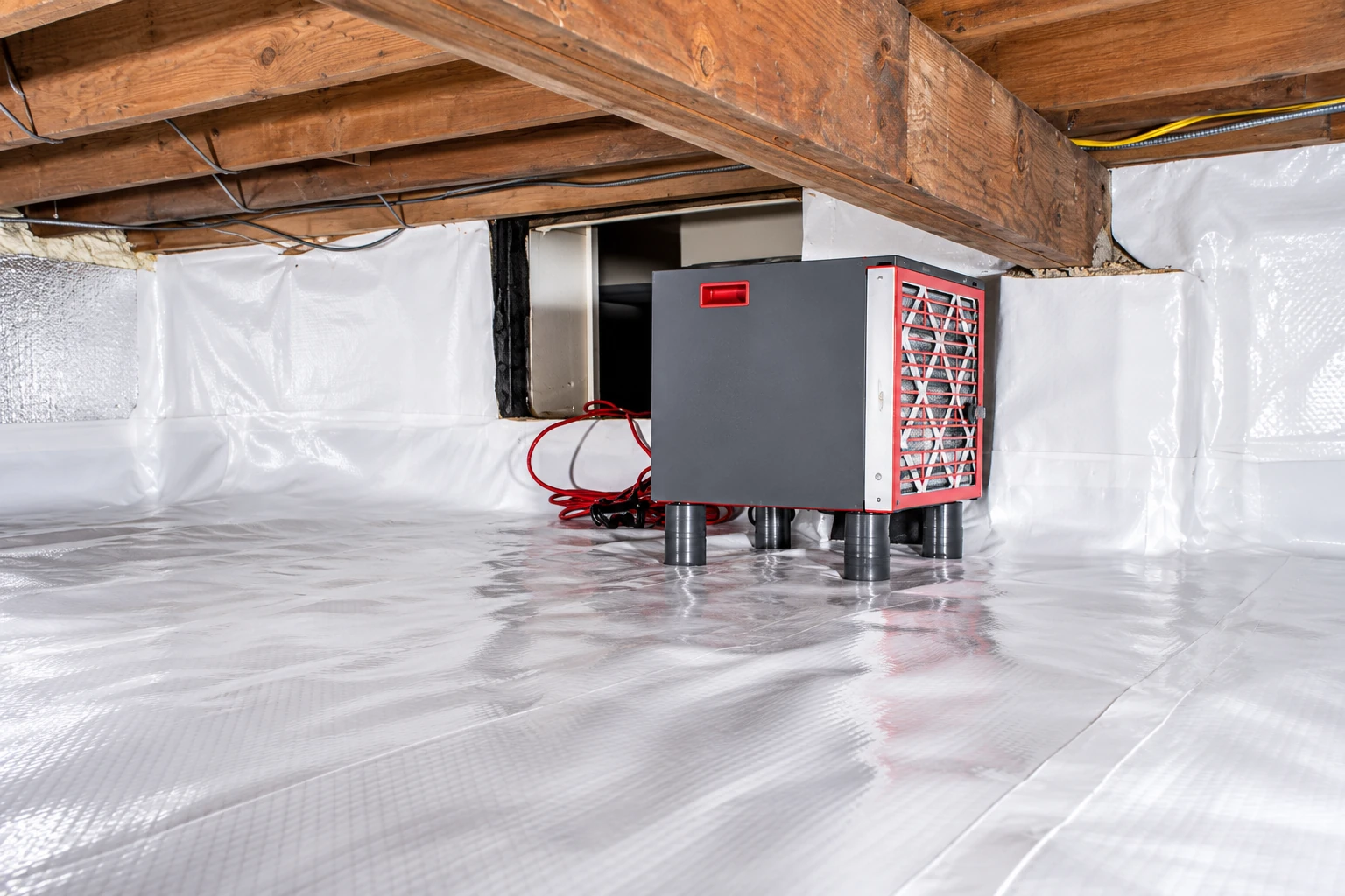 Crawl space with a white vapor barrier covering the floor and walls, beside a gray dehumidifier on short supports.