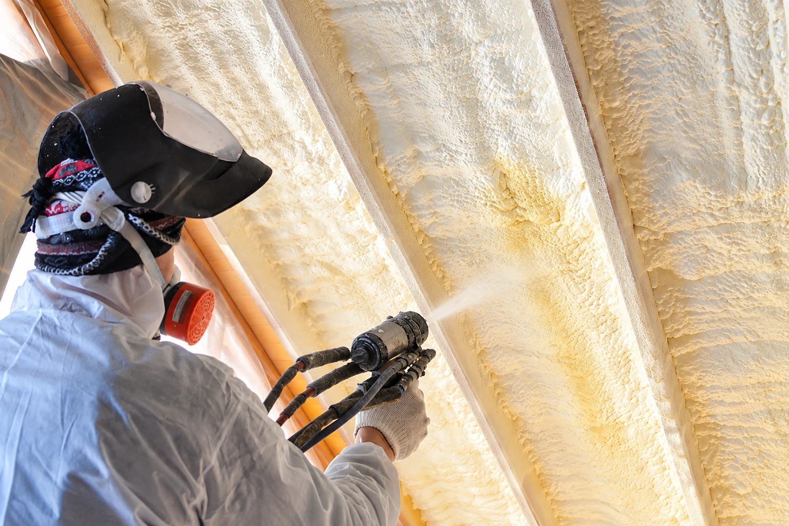 A worker in protective gear sprays foam insulation between exposed roof rafters.