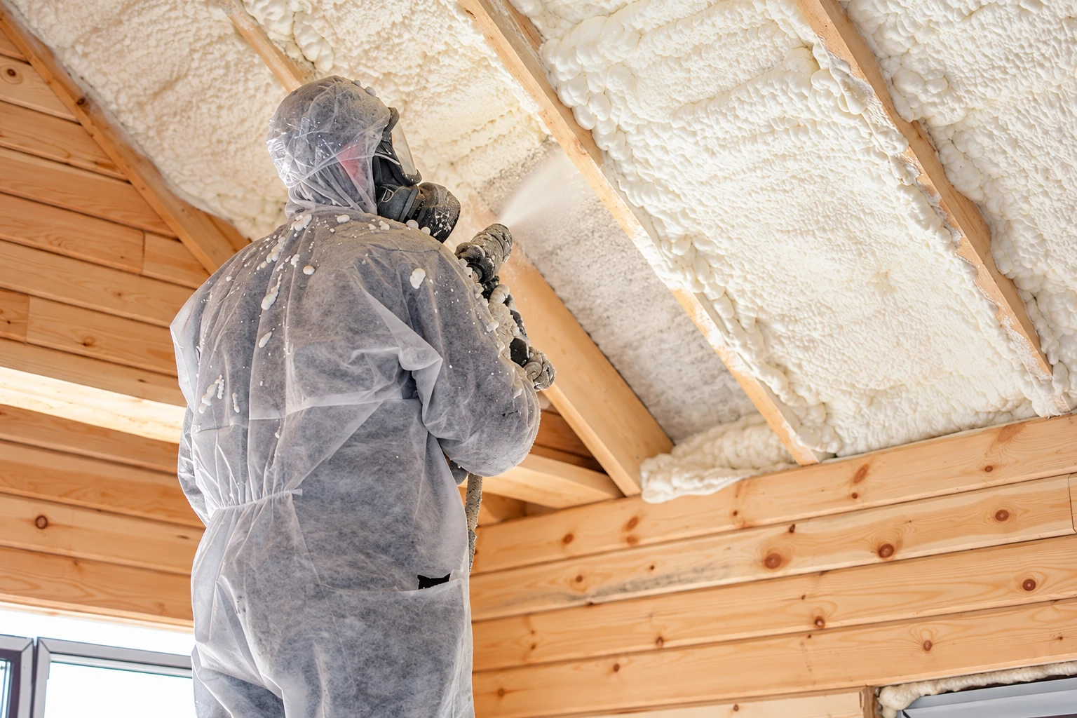 Insulation contractor in protective gear sprays white foam onto the underside of a wooden roof.